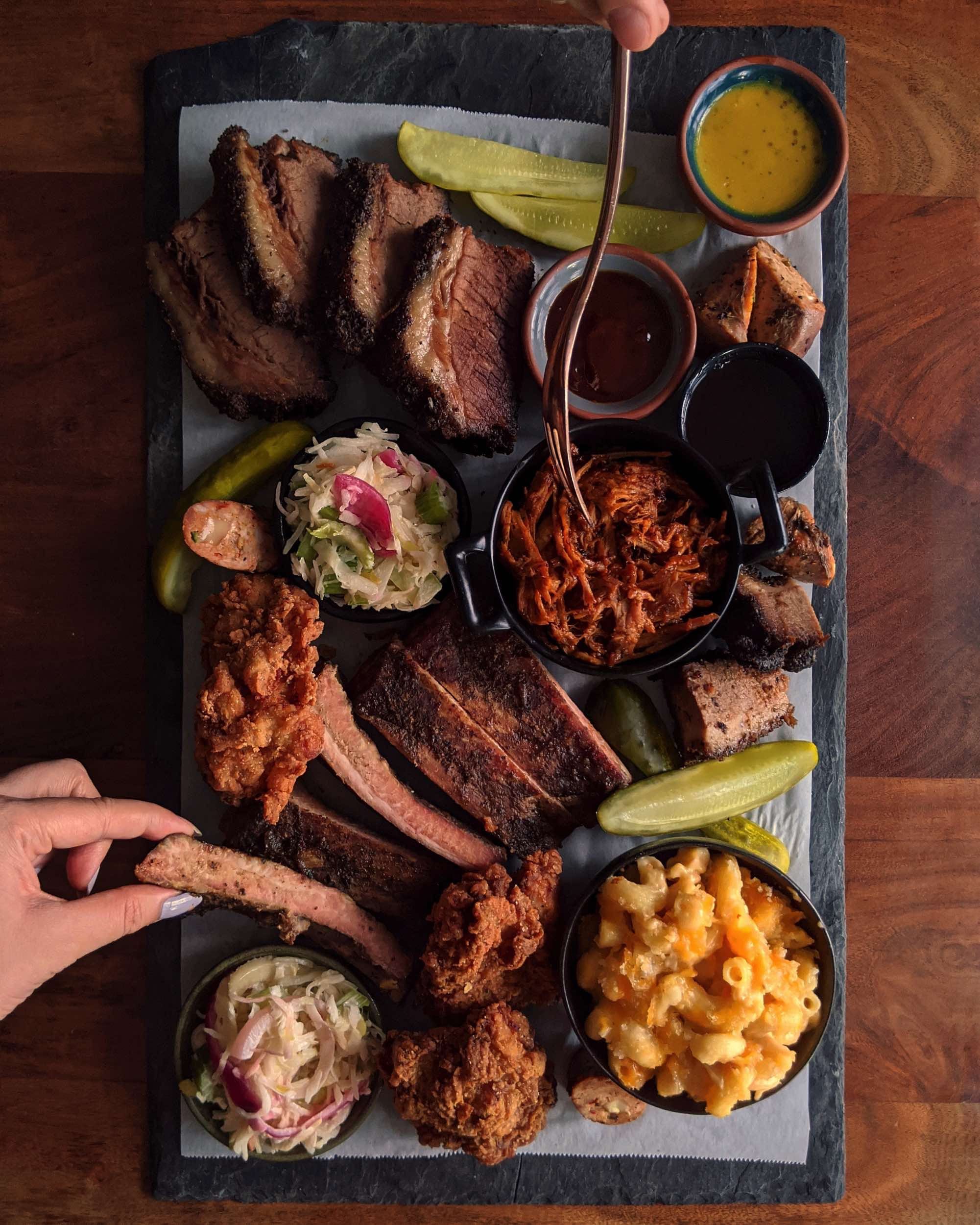 A Carbon Bar BBQ platter with sliced brisket, glossy ribs, crisp fried chicken, pickles, and house sauce